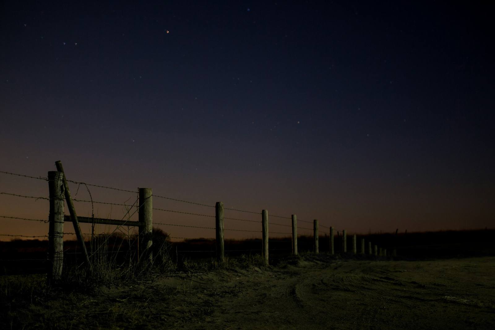 Barbed wire fence line along a dirt field road under a dark night sky.