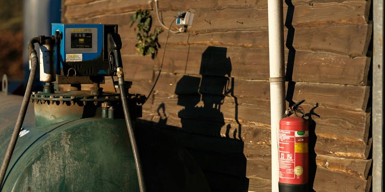 Fuel pump and hose on top of a green farm diesel tank beside a wooden shed wall in low sun.