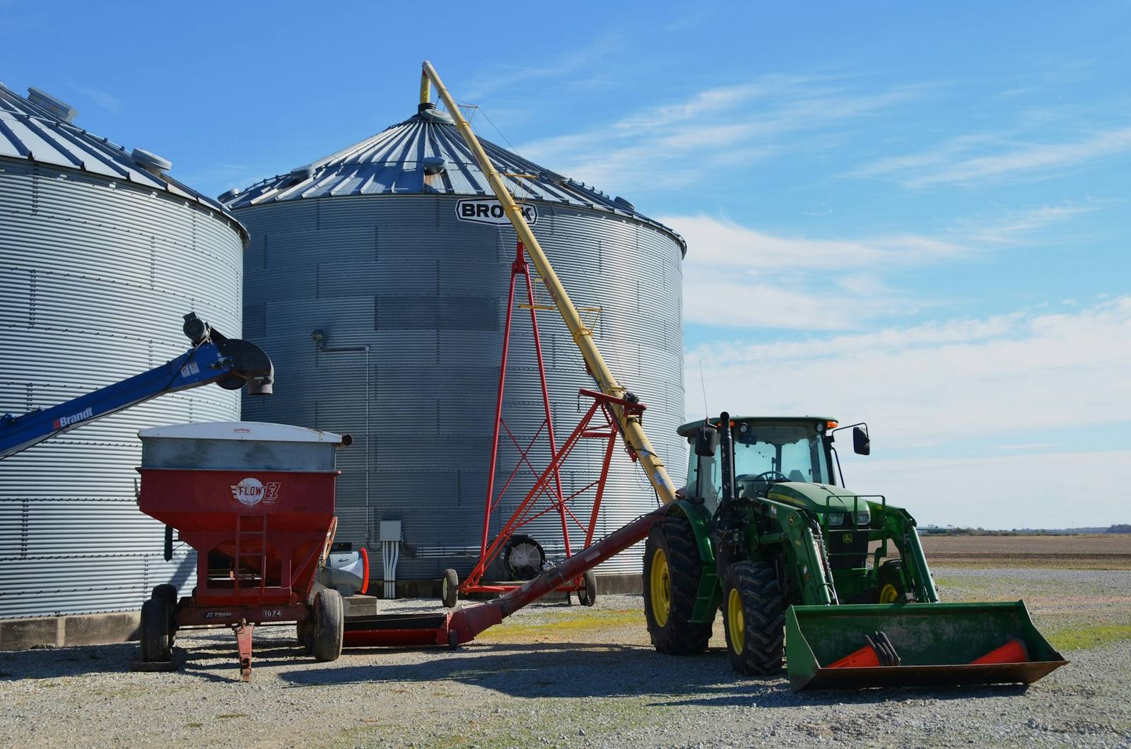 Two corrugated steel grain bins with a red gravity wagon, an auger, and a green loader tractor parked on gravel.