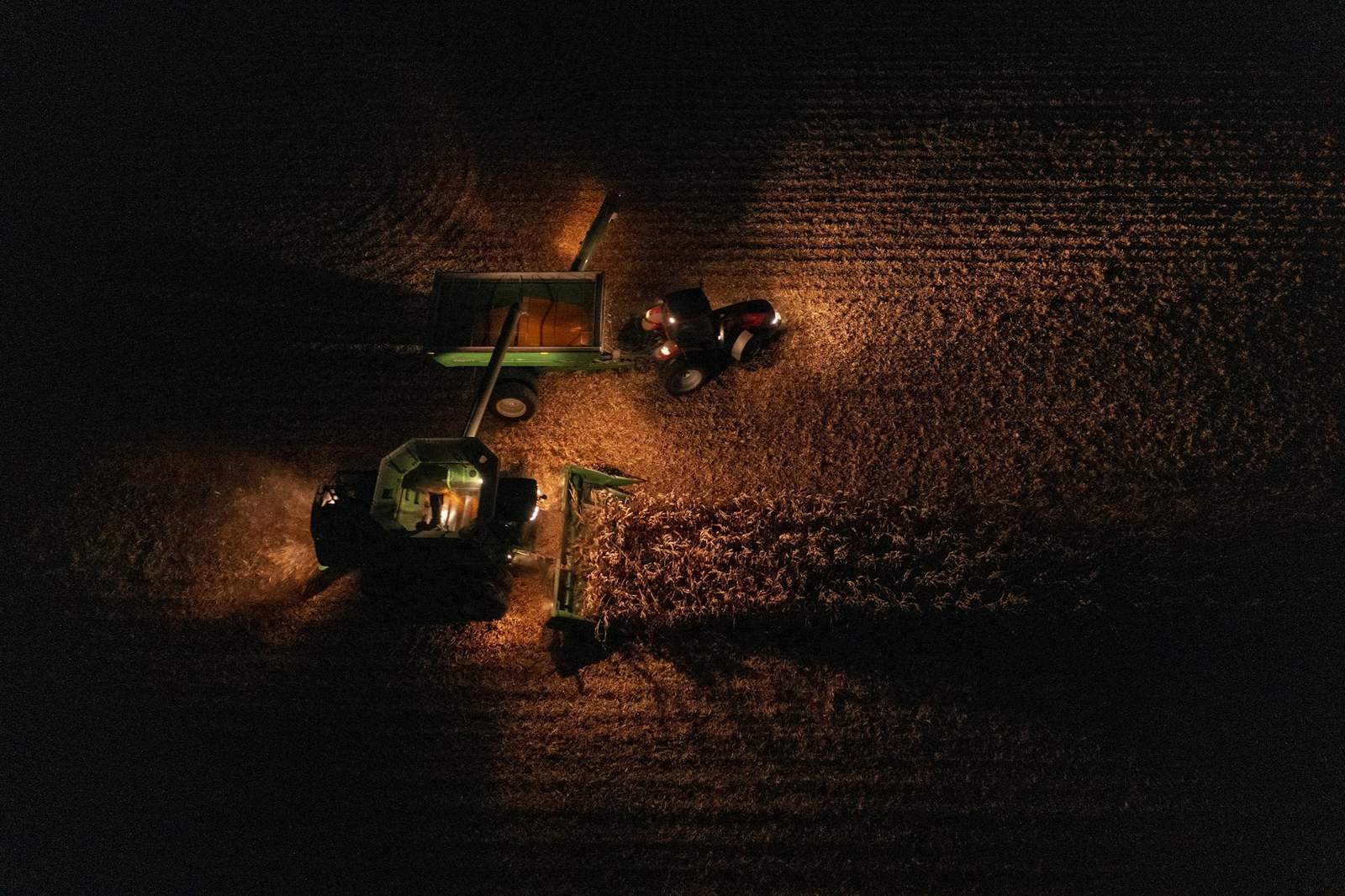 Aerial view at night of a combine unloading corn into a grain cart pulled by a tractor, lit by their work lights, with a dashed GPS boundary drawn over the field.