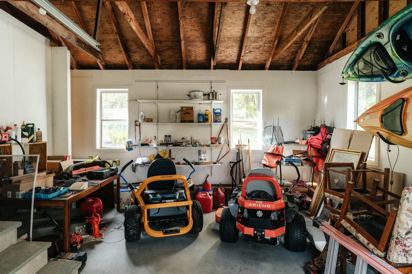 Crowded garage with two riding mowers, red gas cans, a workbench, stacked wooden chairs, and kayaks on the wall