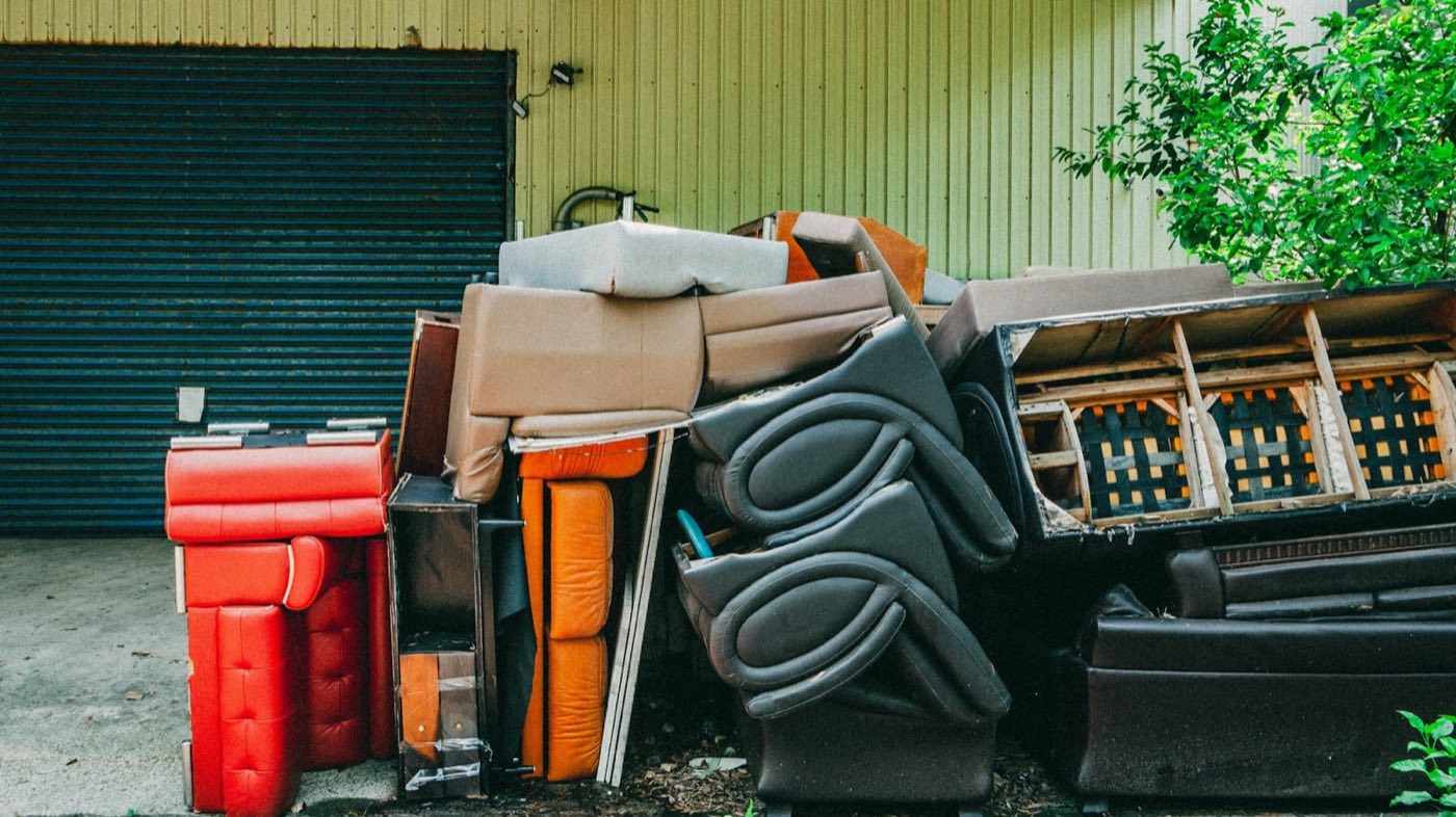 Worn sofas, recliners, and a bed frame stacked against a metal building