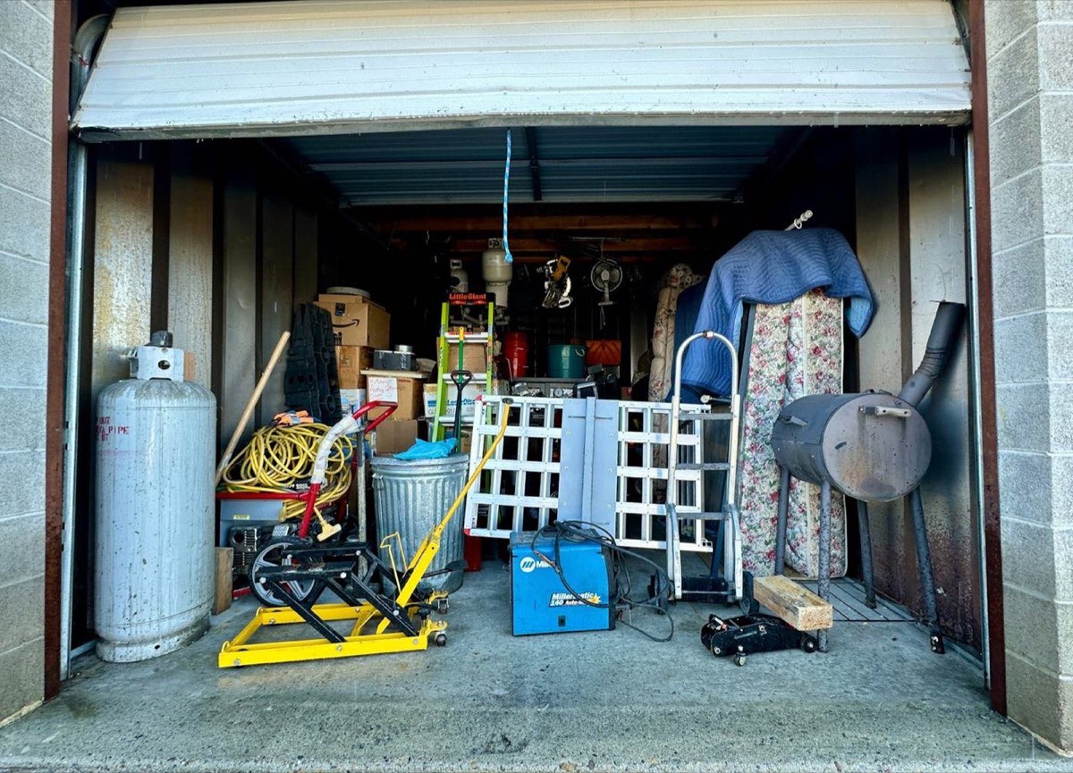 Open storage unit holding a propane tank, a mattress, a smoker grill, a hand truck, and tools