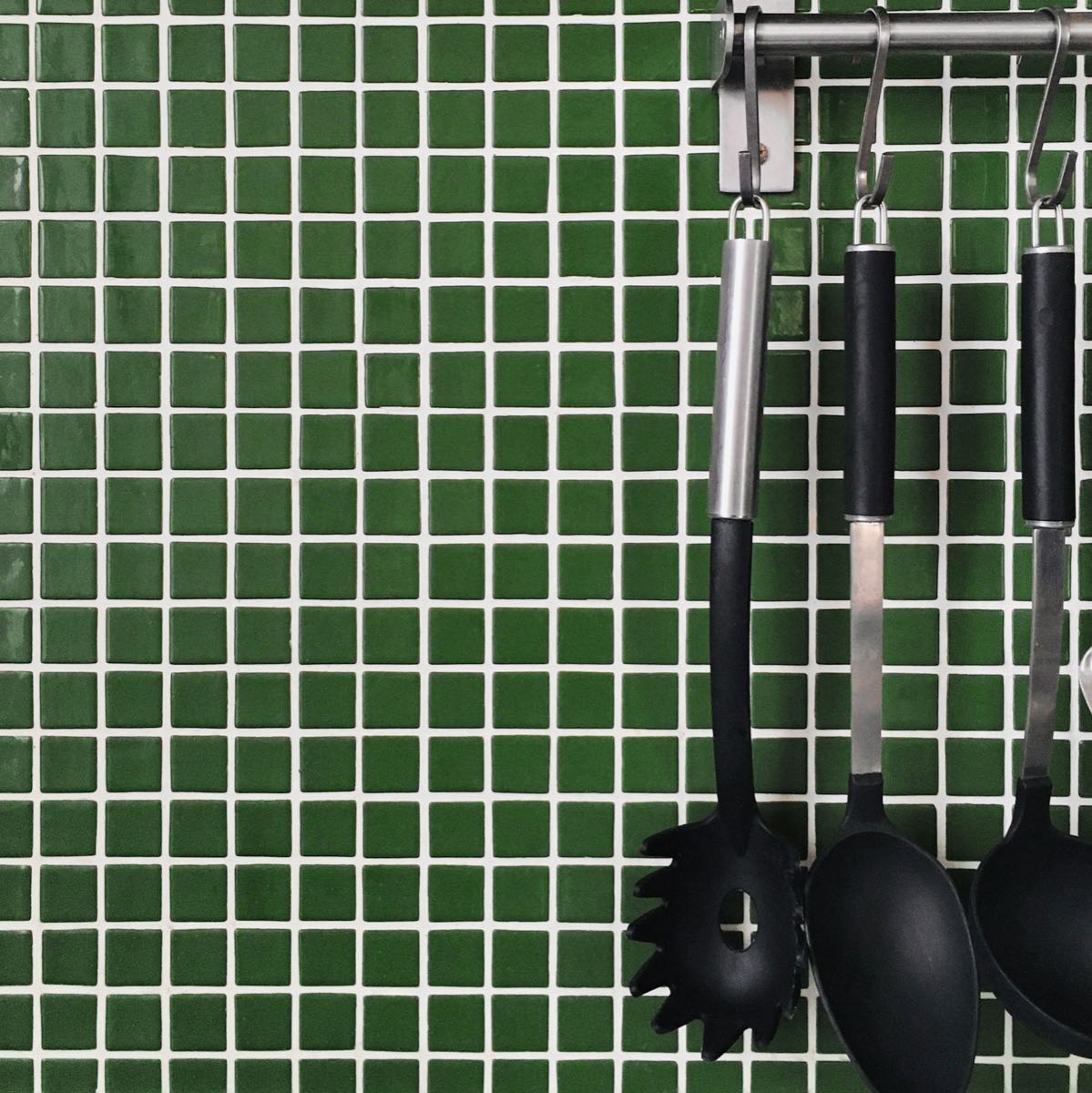 Wall of small dark green square tiles with white grout, with black kitchen utensils hanging from a steel rail