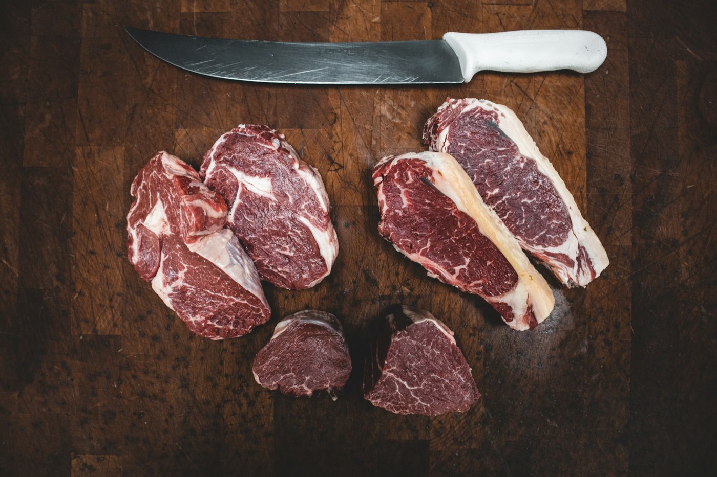 Raw ribeyes, strip steaks and filets on a scarred butcher block beside a butcher knife