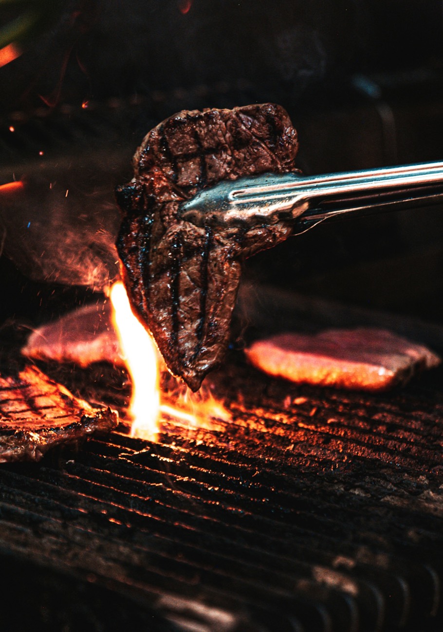 Tongs lifting a grilled steak over open flame