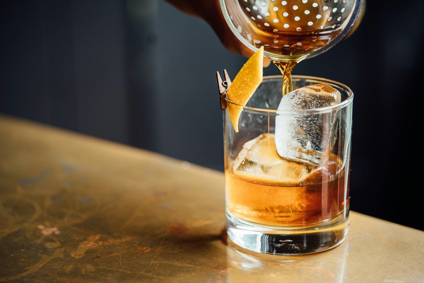 A bartender straining an old fashioned over a large ice cube, orange peel clipped to the glass